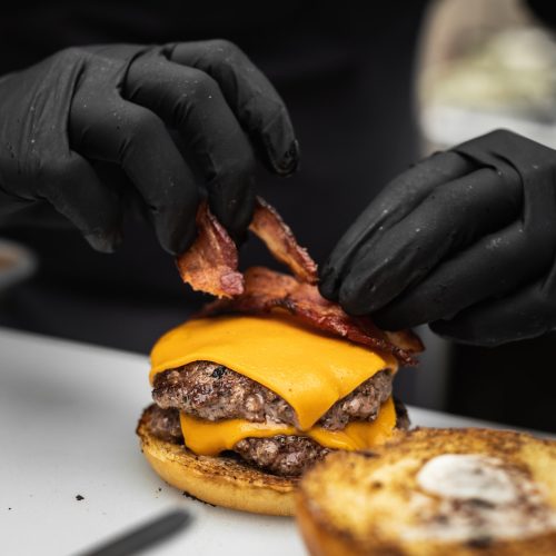 A close-up shot of a cook placing fried bacon over a slice of cheese while layering a burger